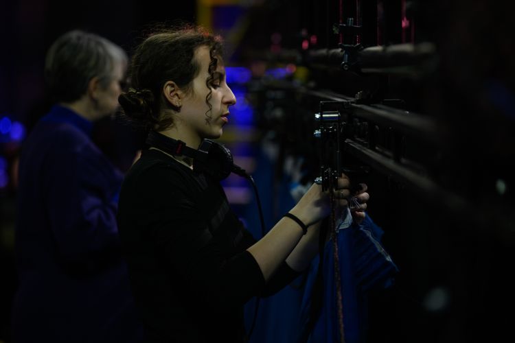 Assistant Production Manager in all black with headphones round their neck fixing something to stage rigging